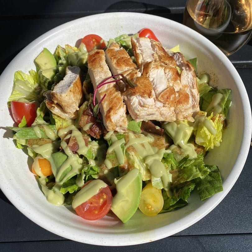 A healthy Good Kitchen salad with grilled chicken, avocado, tomatoes, and green dressing in a white bowl.