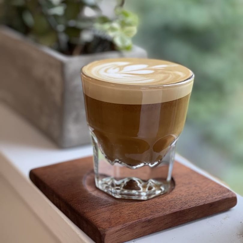 A latte with intricate foam art sits on a wooden coaster by a window at Good Kitchen.