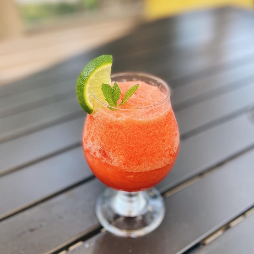 A vibrant red frozen drink, garnished with a lime wedge and mint, sits on a dark wooden table.