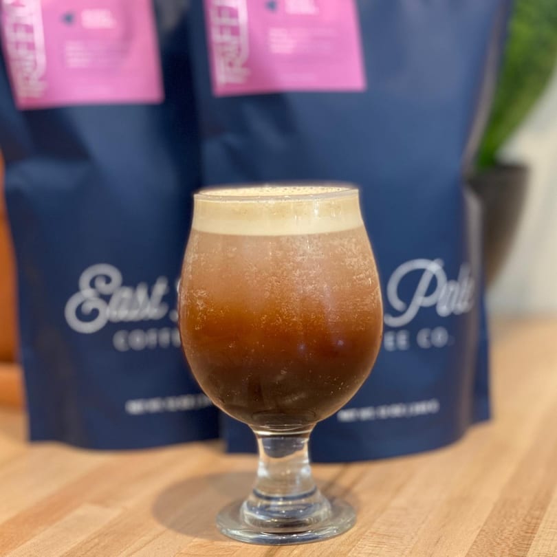 A glass of dark iced coffee with foam on top, with two blue coffee bags in the background.